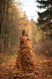 Portrait of young woman covered with leaves standing at forest during autumn