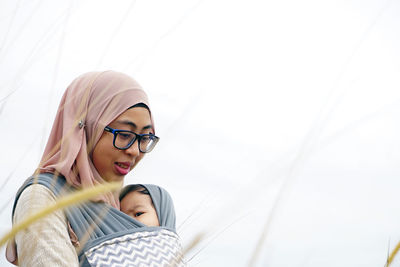 Mother wearing hijab carrying daughter against sky