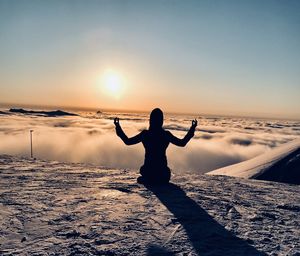 Rear view of woman with arms outstretched meditating while sitting on land against sky during sunset