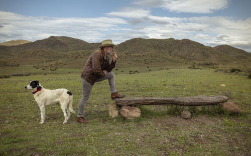 Adult man in cowboy hat and his dog standing on field against sky