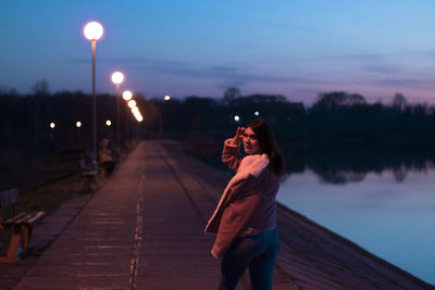 Woman standing on illuminated street light against sky at sunset