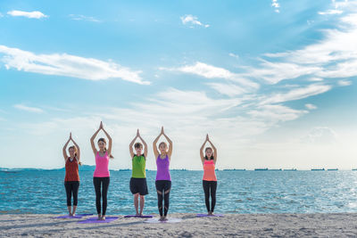 Young friends meditating in prayer position while standing on shore at beach during sunny day