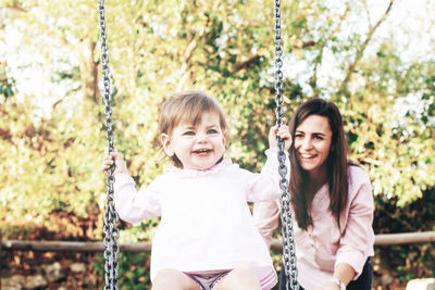 Mother pushing cute daughter swinging against trees in park