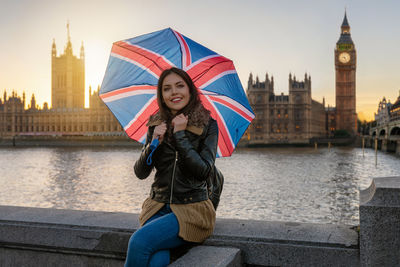 Smiling woman with umbrella sitting on retaining wall against big ben