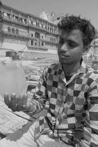 Young man holding plastic container while sitting in city during sunny day