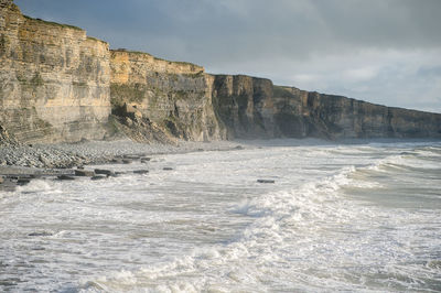 Scenic view of sea waves splashing on cliff against cloudy sky