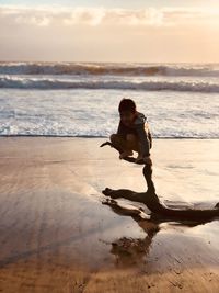Full length of boy sitting on driftwood at beach against sky during sunset