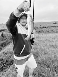 Rear view of young man standing on field with a fishing rod holding a bass fish.