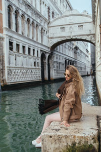 Young woman sitting at canal against buildings