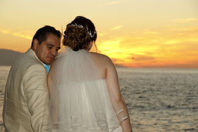 Newlywed couple sitting against sea during sunset