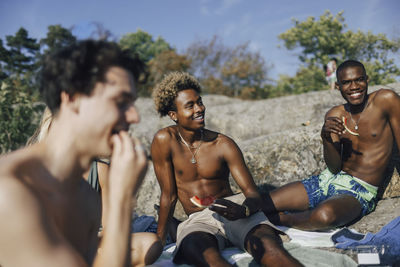 Smiling shirtless male friends eating watermelon while spending weekend with friends
