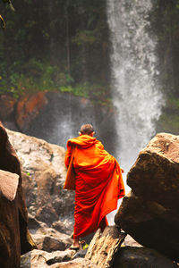 Rear view of monk standing on rock against waterfall