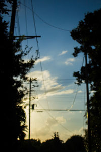 Silhouette trees and electricity pylon against cloudy sky