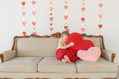 Young blond girl on couch hugging big red plush pillow indoors couch