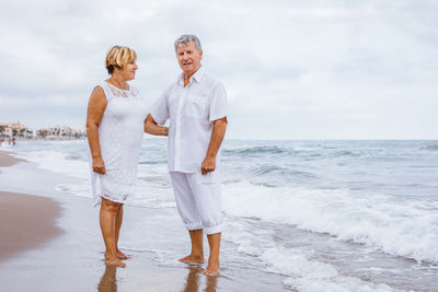 Portrait of romantic senior couple standing at beach against sky
