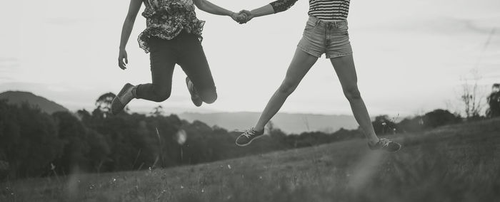 Low section of friends jumping over field against sky