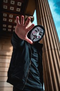 Low angle portrait of man wearing mask while gesturing against column