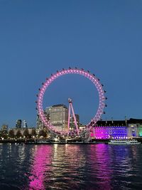 Illuminated ferris wheel by river against sky