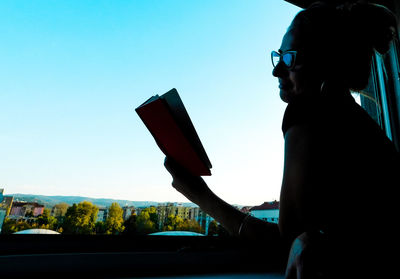 Side view of woman sitting on car against blue sky