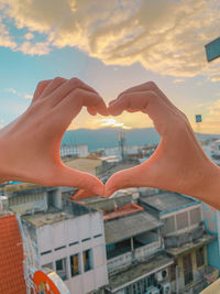 Cropped image of hand holding heart shape against buildings in city during sunset