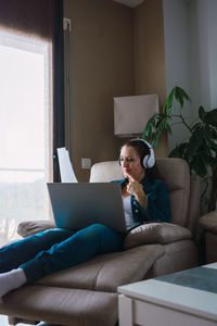 Mature woman listening to music in headphones and reading paper while sitting in armchair and doing remote job on laptop near window at home