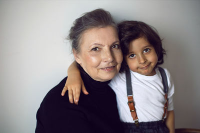 Grandmother with a four-year-old grandson with long hair stands against a white wall with a chair