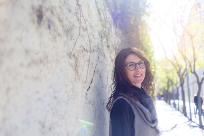 Smiling woman with eyeglasses leaning on wall, looking away