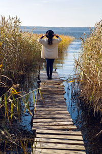 Woman in a black hat and sweater stands on the old pier to the bushes by the lake