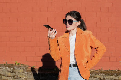 A young brunette in an orange jacket records an audio message on a smartphone