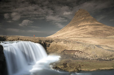 Distant view of man standing by waterfall and mountain against cloudy sky