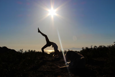 Silhouette woman by dog practicing yoga on cliff against sky during sunset