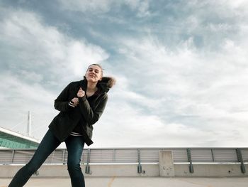 Low angle view of smiling young woman standing on bridge against sky