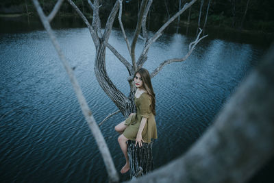 Portrait of young woman sitting over lake on bare tree