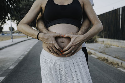 Midsection of pregnant couple making heart shape over abdomen while standing on road