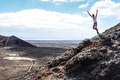 Man with arms raised jumping from rock formation against cloudy sky