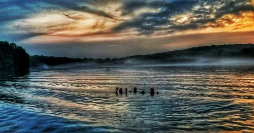 Silhouette people swimming in sea against sky during sunset