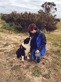 Full length of young man with dog crouching on field against cloudy sky