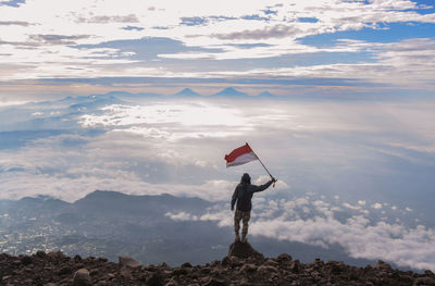 Rear view of man holding flag while standing on rock against sky