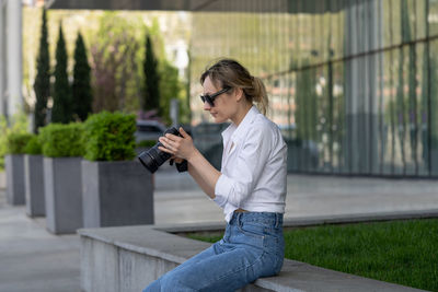 Focused young woman sitting on bench in spring park under tree outdoors resting using camera