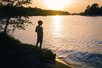 Rear view of man standing by lake against sky during sunset