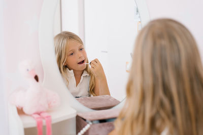 Pretty child girl paints her lips with glitter or balm in front of a mirror in her room