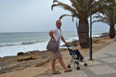 Portrait of man standing with baby stroller on promenade against sky