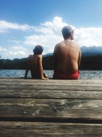 Rear view of shirtless man and boy sitting by lake against sky