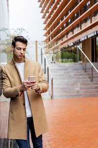 Young man using phone while standing by wall