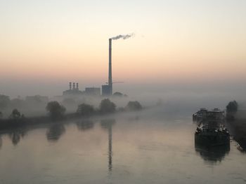 Smoke emitting from factory by river during foggy weather