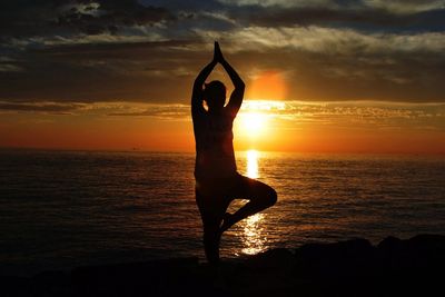 Silhouette of woman practicing yoga by sea against cloudy sky at sunset
