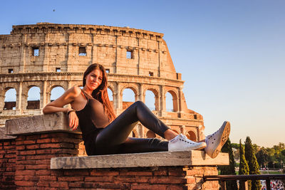 Young woman sitting on historical building against sky