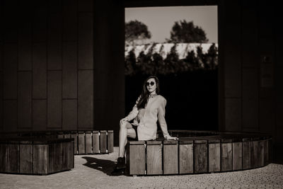 Full length of fashionable woman wearing sunglasses while sitting on wooden seats during sunny day