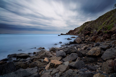 Scenic view of mountain at sea shore against cloudy sky during sunset