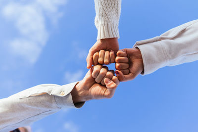 From below of faceless close friends bumping fists against clear blue sky in sunlight